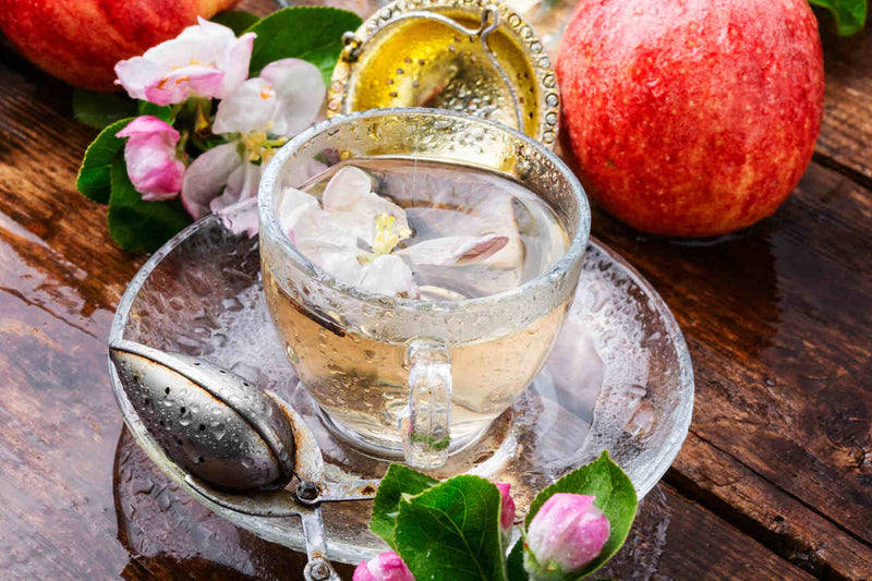 Ahşap bir masa üzerinde şeffaf bir fincan, içerisinde elma çayı, çiçek yaprakları ve yanında kırmızı elmalar